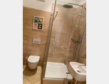 Modernes Badezimmer mit Wand- und Bodenfliesen in Beige, einer eckigen Duschkabine mit Regenduschkopf, wandhängender Toilette und kleinem Waschbecken. An der Wand hängt ein gerahmtes Bild mit geometrischen Formen und dem Wort „LOVE“.