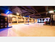 Ein moderner Veranstaltungsraum mit glänzenden Spiegelpfeilern, hängenden runden Leuchtelementen und einer offenen Fläche mit hellen Fliesen. Im Hintergrund befinden sich Sitzbereiche mit Holzelementen und große Fenster mit schweren Vorhängen.