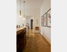 Minimalistisch gestalteter Innenraum mit hellen Wänden, einem Parkettboden im Fischgrätmuster, einer Kücheninsel aus Holz und zwei hängenden Lampen mit goldenen Fassungen. An der rechten Wand hängen mehrere gerahmte Bilder, und im Hintergrund steht ein schlichter Stuhl.