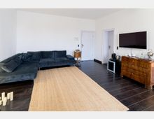Helles Wohnzimmer mit weiß gestrichenen Wänden, dunklem Holzboden und großem beigem Teppich. Links steht ein großes dunkelgraues Ecksofa, rechts eine Holzkommode mit montiertem Flachbildfernseher darüber sowie Kaffee-/Teestation neben zwei Türen.