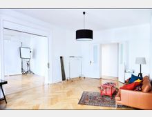 Helles Wohnzimmer mit Parkettboden und weißen Wänden, eingerichtet mit einem braunen Ledersofa, bunten Kissen, einem roten Couchtisch und einem mehrfarbigen Teppich. Ein angrenzender Raum mit Fotostudio-Equipment ist durch eine breite, offene Tür sichtbar.