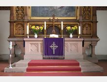 Steinaltar in einer Kirche mit einem violetten Altartuch, darauf ein Kreuzsymbol. Der Altar ist mit Kerzen, einem Kruzifix, gelben Blumen in weißen Vasen und einer geöffneten Bibel dekoriert. Im Hintergrund ein kunstvoll geschnitztes hölzernes Retabel und ein Gemälde.