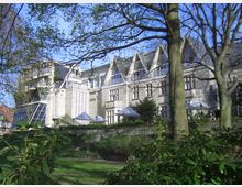 Steinernes Gebäude mit spitz zulaufenden Giebeln und großen Fenstern, eingebettet in eine parkähnliche Umgebung mit Bäumen und Sträuchern; im Vordergrund Sonnenschirme, die auf einen Außenbereich hinweisen.