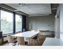 Moderner Besprechungsraum mit schlichten Holzmöbeln, weißen Tischen und großen Fenstern, die den Blick auf Bäume und umliegende Gebäude bieten. Die Wände sind aus Sichtbeton, der Raum hat einen Holzfußboden und eine abgehängte Decke mit integrierter Beleuchtung.