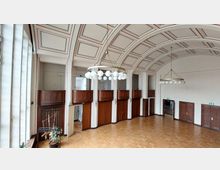 Großer Innenraum mit Holztäfelungen an den Wänden, hohen Fenstern auf der linken Seite und einer gewölbten Decke mit dekorativen roten und grauen Linien. Zwei große, ringförmige Kronleuchter mit kugelförmigen Lampen hängen von der Decke, und auf dem Boden liegt ein heller Holzparkettboden.