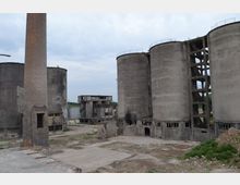 Industriegelände mit verfallenen Gebäuden, darunter mehrere große Betonsilos mit beschädigten Fassaden und ein hoher gemauerter Schornstein. Der Boden ist uneben und zeigt Spuren von Zerstörung, umgeben von einigen Grünflächen im Hintergrund.