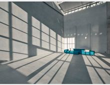 Minimalistischer Innenraum mit hohen, grauen Wänden und einem großen Oberlicht, das Schattenspiele von Fensterrahmen auf dem Boden und den Wänden erzeugt. In der Ecke steht ein niedriges, türkisfarbenes Sofa mit weißen Kissen.