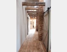 Ein schmaler Flur mit Holzdielenboden, weißen Wänden und freiliegenden Holzbalken an der Decke. Rechts ist eine unverputzte Ziegelwand, daneben eine Schiebetür aus Holz; im Hintergrund ist ein heller Raum sichtbar.