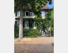 Zweistöckiges Wohnhaus mit weißer Fassade, schwarzen Fensterläden und einem roten Ziegeldach, teilweise bewachsen mit grünem Efeu. Im Vordergrund befinden sich ein Gehweg, ein Metallzaun mit Tor und ein Briefkasten, daneben ein großer Straßenbaum.