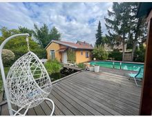 Garten mit einer Holzterrasse, auf der sich ein weißer Hängesessel und ein türkisfarbener Liegestuhl befinden. Im Hintergrund ein Pool, ein gelbes Gartenhaus mit rotem Dach, ein kleiner Teich mit Pflanzen sowie hohe Bäume und eine Hecke als Begrenzung.