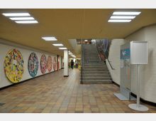 Ein heller, gefliester Innenbereich mit rechteckigen Deckenleuchten, einer Treppe rechts, die nach oben führt, und runden, bunt gestalteten Tafeln an der linken Wand. Im Hintergrund sind ein Gang mit Menschen sowie ein Informationsschild sichtbar.