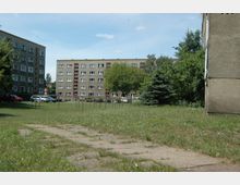 Freifläche mit Gras und Gehwegplatten in einem Wohngebiet, umgeben von mehreren mehrstöckigen Wohnblocks mit beige-grauen Fassaden und symmetrisch angeordneten Fenstern; im Hintergrund Parkplätze und Bäume.