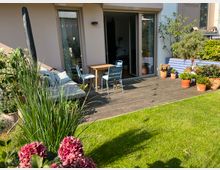 Wohnterrasse mit Holzfußboden, umgeben von grüner Rasenfläche und blühenden Pflanzen in Töpfen. Im Hintergrund sind eine geöffnete Glastür, ein Tisch mit Stühlen und eine Liege zu sehen; daneben stehen Hochbeete und weitere Topfpflanzen.