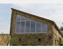 Historisches Gebäude aus Backstein mit einem asymmetrischen Satteldach und großen, modernen Fenstern im oberen Bereich. Die Fassade weist Rundbogenfenster und dekorative Mauerwerksdetails auf, umgeben von Bäumen und einer blauen Himmelsperspektive.