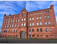 Backstein-Gebäude mit symmetrischer Fassade und mehreren Etagen, geprägt durch hohe Fensterbögen und dekorative Zierelemente. Der Eingangsbereich umfasst ein großes Tor, und die Umgebung besteht aus einem gepflasterten Hof unter blauem Himmel.