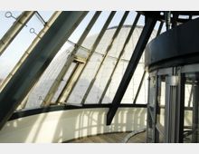 Innenansicht einer modernen Kuppelkonstruktion mit einer Kombination aus Glasfenstern und Metallstreben. Im Vordergrund ist ein runder Glasaufzug zu sehen, während die gewölbte, metallisch verkleidete Außenkuppel im Hintergrund sichtbar ist.