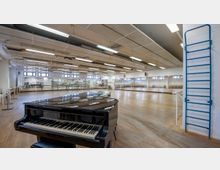 Großer Tanz- oder Übungsraum mit Holzboden, bodentiefen Spiegeln an einer Wand und Übungsstangen entlang der Seiten. Im Vordergrund steht ein schwarzer Flügel, und an der rechten Wand ist eine blaue Sprossenwand angebracht.