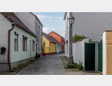 Schmale Kopfsteinpflaster-Gasse mit kleinen, farbigen Wohnhäusern und roten Ziegeldächern; links und rechts stehen Häuser in Pastell- und Erdtönen. Eine Laterne und ein grünes Gartentor säumen den Weg.