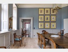 Esszimmer in einem historischen Gebäude mit hellblauen Wänden, Holzfußboden und einem langen Esstisch mit Holzstühlen. An der Wand hängen gerahmte Drucke, und im Hintergrund ist ein Flur mit schwarz-weißem Schachbrettboden sichtbar.