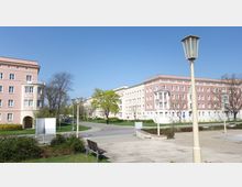 Wohnsiedlung mit mehrstöckigen, hellrosa und cremefarbenen Gebäuden im Stil des sozialistischen Klassizismus, umgeben von Bäumen, Gehwegen und einer gepflasterten Platzfläche mit Parkbänken und Laternen. Im Hintergrund erstrecken sich Grünflächen und eine kleine Straße.