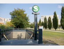 Eingang zu einer S-Bahn-Station mit einem runden „S“-Logo und der Beschriftung „Anhalter Bahnhof“ auf einer Säule. Im Hintergrund sind Wohngebäude, Bäume und eine Grünfläche mit einem Tennisplatz sichtbar.
