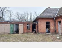 Ein verfallenes Gebäude aus rotem Ziegelstein mit mehreren beschädigten Türen und Fenstern. Im Vordergrund befindet sich eine unregelmäßige Pflasterfläche sowie kahler Boden, im Hintergrund stehen unbelaubte Bäume.