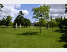 Weitläufiger Park mit grüner Wiese und verstreuten Bäumen, im Hintergrund das Reichstagsgebäude mit seiner gläsernen Kuppel sichtbar. Vereinzelte Steinskulpturen stehen zwischen den Bäumen, der Himmel ist teils bewölkt.