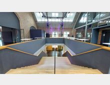 Innenansicht eines modernen Event- oder Konferenzraums in einem Gebäude mit industriellem Stil. Sichtbar sind eine breite Holztreppe, Wände aus Backstein und Glas sowie ein hoher Raum mit Oberlichtern und Stahlträgern.