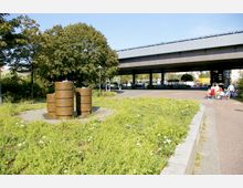 Eine Grünfläche mit einem kleinen Springbrunnen aus gestapelten, bronzefarbenen Zylindern im Vordergrund, umgeben von wild wachsenden Blumen und Wiesen. Im Hintergrund verläuft eine erhöhte Straßenbrücke, unter der sich eine gepflasterte Fläche und Gehwege mit Passanten befinden.
