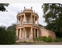 Runder, zweigeschossiger historischer Pavillon im neoklassizistischen Stil mit Säulen und Verzierungen, umgeben von Bäumen und Sträuchern in einer parkähnlichen Landschaft. Eine geschwungene Freitreppe führt seitlich zum oberen Bereich des Gebäudes.
