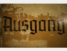 Beige Wand mit abgeschälter Farbe, auf der in schwarzer Frakturschrift das Wort „Ausgang“ steht; der Hintergrund wirkt alt und abgenutzt.