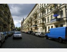 Straßenszene in einer Wohngegend mit mehrstöckigen Altbauten im Gründerzeitstil auf beiden Seiten, Balkonen mit Blumen und parkenden Autos entlang der gepflasterten Straße. Im Hintergrund ragt die Spitze eines Backsteinturms empor, unter einem wolkigen Himmel mit etwas Licht.