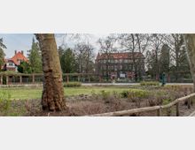 Historisches Backsteingebäude mit rotem Satteldach, umgeben von Bäumen und einem Zaun aus Ziegelpfeilern; im Vordergrund ein angelegter Gartenbereich mit Wiese, Sträuchern und einem Gehweg aus Holzstämmen.