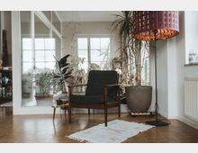 Ein heller Raum mit einem Holzfußboden, in dem ein grüner Sessel mit Holzrahmen, eine große Topfpflanze und eine Stehlampe mit violettem Lampenschirm stehen. Im Hintergrund sind große Fenster und Glastüren, die zu einem weiteren Zimmer mit Pflanzen führen.