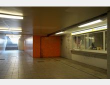 Unterführung mit gefliestem Boden, orangefarbenen Fliesenwänden und Deckenbeleuchtung. Rechts befindet sich ein Fahrkartenschalter mit der Aufschrift „Fahrscheine und mehr“, links führen Treppen ins Freie.