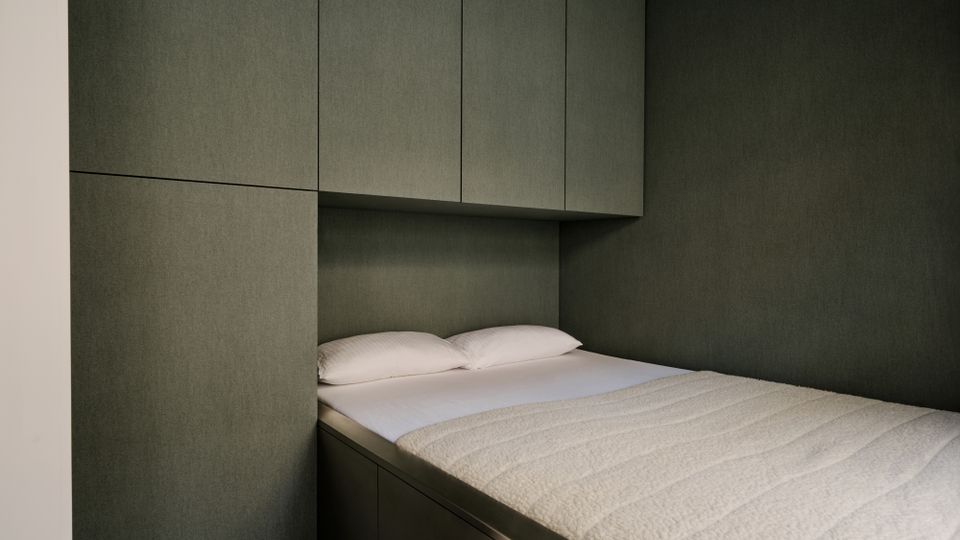 Schmaler Schlafbereich mit einem eingebauten Bett mit weißer Bettwäsche, umgeben von dunkelgrauen Wand- und Schrankpaneelen. Über dem Kopfteil befinden sich bündig integrierte Hängeschränke, das Bettgestell wirkt als Einbaumöbel mit Stauraum.