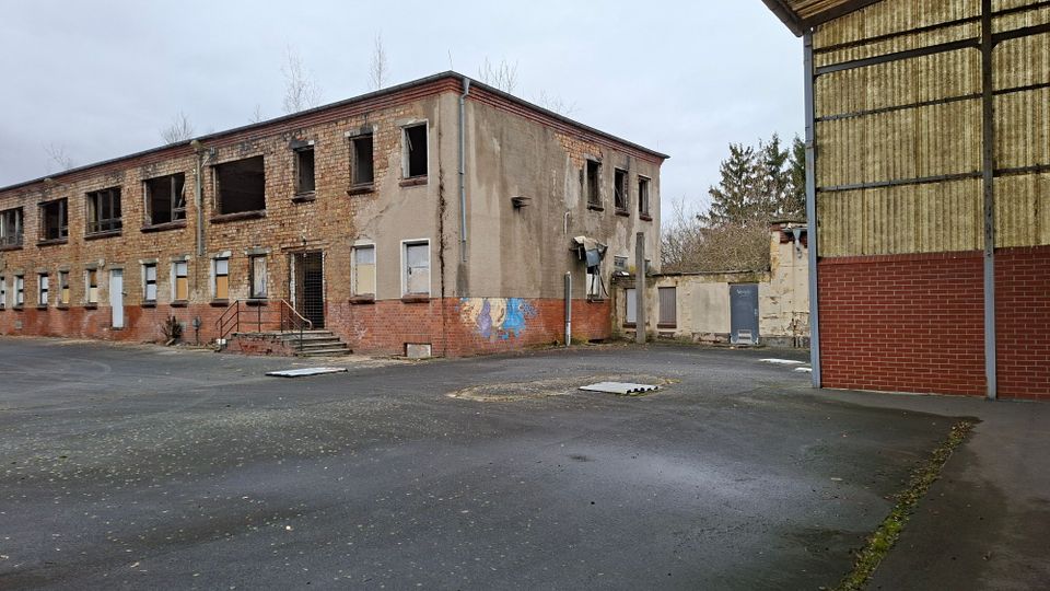 Verlassener Industriehof mit einem zweigeschossigen, teils verputzten Backsteingebäude mit zerbrochenen Fenstern und zugemauerten Öffnungen sowie einem Eingang mit Stufen. Rechts steht eine große Hallenwand aus Wellplatten und rotem Mauerwerk, davor eine breite asphaltierte Freifläche.