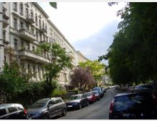 Straße mit geparkten Autos, flankiert von historischen Wohngebäuden mit dekorativen Fassaden und Balkonen auf der linken Seite sowie dichtem Baumbewuchs auf der rechten Seite. Die Umgebung wirkt ruhig und von Grünflächen geprägt.