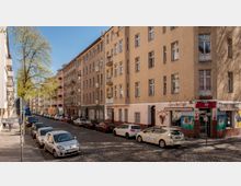 Straßenszene in einer Wohngegend mit mehrstöckigen Mietshäusern im Altbaustil, einer Baumreihe entlang der Straße und parkenden Autos. Im Erdgeschoss eines Gebäudes ist ein Café mit bunter Fassadengestaltung zu sehen, daneben ein Straßenschild mit der Aufschrift „Wipperstr.“.