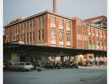 Backsteinfassade eines Industriegebäudes mit großen Fenstern und einem Schornstein im Hintergrund. Im Vordergrund stehen Motorräder und ein geparkter Oldtimer auf einem gepflasterten Hof, umgeben von einzelnen Personen.