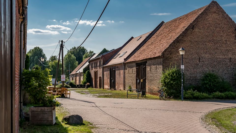 Dorfstraße mit gepflasterter Fahrbahn und mehreren niedrigen Backsteingebäuden mit Satteldächern, darunter ein großes Scheunenhaus mit Holztoren. Entlang der Straße stehen Laternen und Stromleitungen, daneben Grünflächen und Büsche am Rand.