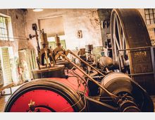 Innenansicht eines historischen Maschinenraums mit einer großen, dampfbetriebenen Maschine. Zu sehen sind ein rotes Kesselgehäuse, Metallventile, Hebel sowie ein großes Schwungrad; im Hintergrund befinden sich alte Wände und Fenster mit Gitterstruktur.