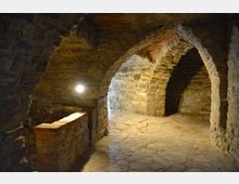 Steinerner Gewölbekeller mit dicken, unregelmäßigen Steinwänden und runden Bögen, die unterschiedliche Bereiche des Raumes abgrenzen. Der Boden ist aus groben Steinplatten, und eine kleine Mauer aus Ziegelsteinen befindet sich im Vordergrund, während eine Lampe die Szene beleuchtet.