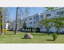 Ein modernes, weißes Wohngebäude mit großen Fenstern und minimalistischer Architektur, umgeben von einer gepflegten Grünfläche mit einzelnen Bäumen und Sträuchern. Im Vordergrund steht ein großer Gedenkstein auf einer Wiese.