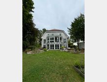 Zweistöckiges Wohnhaus mit hellem Putz, großen Glasfenstern und einem Balkon im oberen Stockwerk, umgeben von einem gepflegten Garten mit Rasenfläche und Bepflanzungen, sowie Bäumen im Hintergrund.