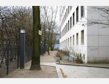 Gehweg zwischen einem modernen Bürogebäude aus hellem Stein mit rechteckigen Fenstern und einem kargen Außenbereich mit Bäumen, niedrigem Gestrüpp, Metallzaun und Wegebeleuchtung. Im Hintergrund ist ein weiteres Gebäude zu sehen.