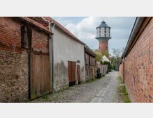 Schmale, gepflasterte Gasse mit alten Ziegel- und Putzgebäuden links und einer rotbraunen Ziegelmauer rechts, im Hintergrund ein markanter Wasserturm mit rundem Ziegelunterbau und hellem Aufsatz.