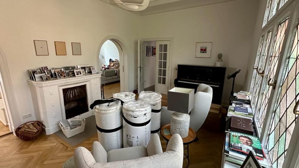 Wohnzimmer mit Holzboden und großen bleiverglasten Fenstern, ausgestattet mit einem weißen Kamin, einem schwarzen Klavier und hellen Polstermöbeln. Ein runder Deckenleuchter hängt zentral, und auf einem Tisch sowie Regalen liegen Zeitschriften und Bücher.