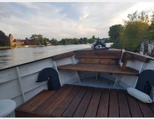 Das Bild zeigt den Bug eines kleinen Boots mit Holzsitzbänken und Boden aus Holzlatten. Im Hintergrund ist ein Fluss zu sehen, gesäumt von Bäumen sowie einem historischen roten Gebäude am Ufer.