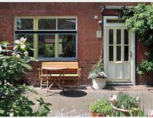 Fassade eines Hauses mit roter Putzwand und gelb-weißer Eingangstür. Davor stehen eine Holz-Sitzbank und ein runder Holztisch, umgeben von Blumentöpfen und Pflanzen in einem kleinen Hofgarten.